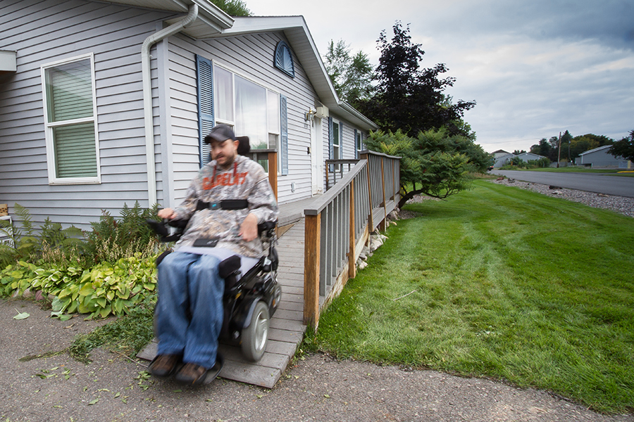 The man drives his power wheelchair off the end of the grey ramp onto the driveway. Towards the street is a green lawn with several manicured bushes near the home.
